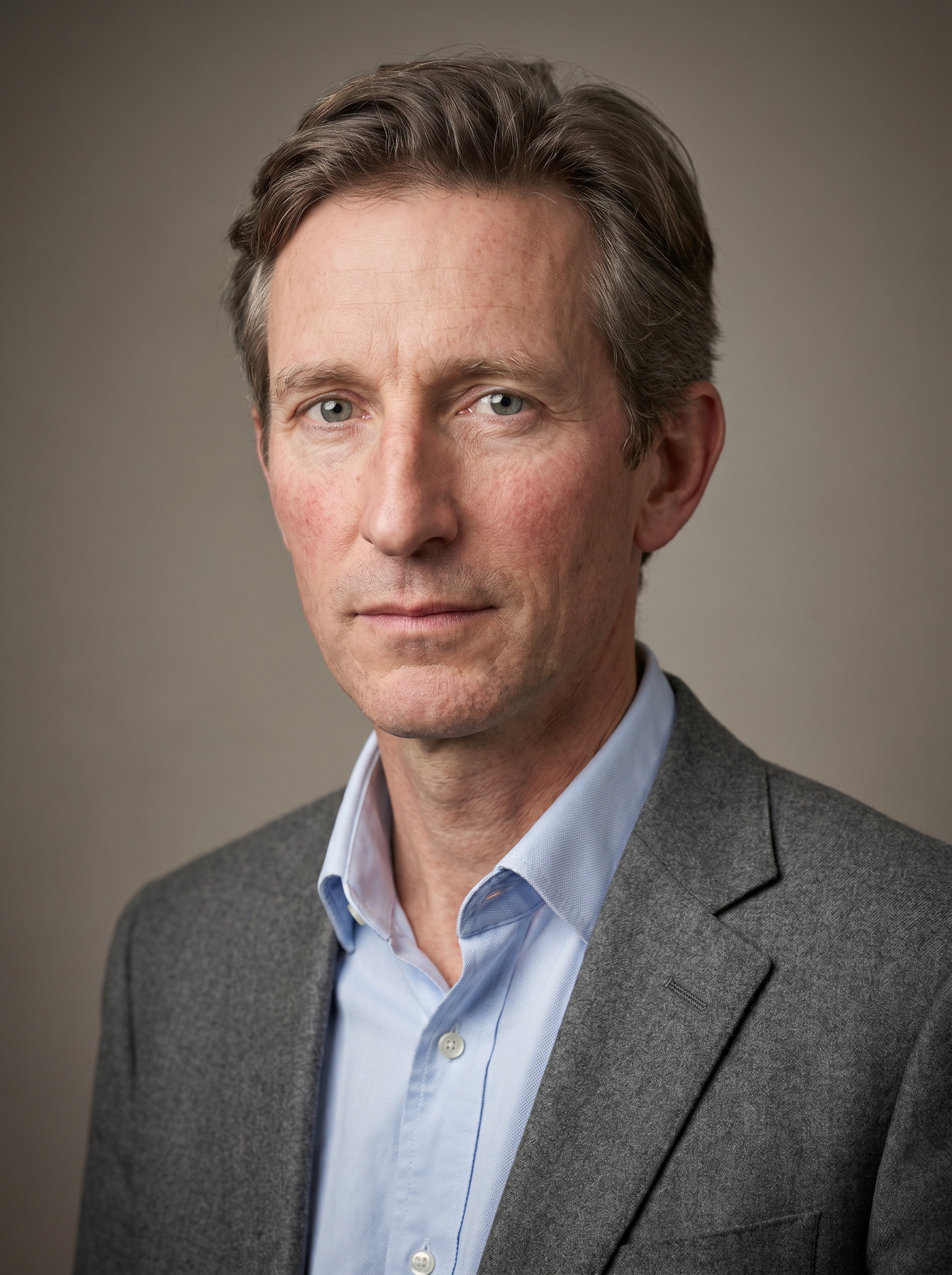 An Irish man in his mid-40s, lean and sharp-featured, with the kind of face that looks lived-in and precisely his age. Fair skin with a light ruddy undertone across the cheeks, faint creasing at the corners of the eyes and along the brow from years of attentive expression. Pale grey-green eyes, alert and steady. His hair is a medium ash-brown, slightly wavy, cut short at the sides and left with a little length on top, threaded with early grey at the temples. Clean-shaven with a narrow jaw and a slim, angular build that suits a man who moves through seminar rooms and accreditation panels with equal ease. He wears a well-fitted mid-grey wool blazer over a tucked pale blue Oxford shirt, collar open by one button, projecting professional warmth without formality. No jewellery beyond a slim brushed-steel watch. Distinguishing feature: a long, prominent aquiline nose with a slight downward curve at the bridge, giving his profile an immediately distinctive, scholarly character that anchors the face across every image.