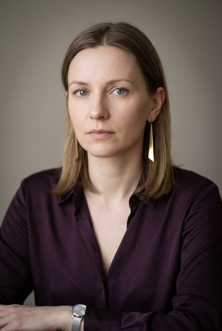 A composed Croatian woman in her late 30s, Zagreb-born and Zurich-based. Fair complexion with cool undertones. Dark-blonde hair worn in a smooth shoulder-length cut, parted in the middle and tucked behind the ears. Pale hazel eyes, neatly defined eyebrows. Slim build with calm posture. A deep aubergine silk blouse tucked into slim mid-grey wool trousers, grey suede ankle boots, a slim silver watch on the left wrist. Distinguishing feature: a single long architectural brass drop earring worn in the left ear only, the right ear bare — a deliberate asymmetric statement piece, geometric and modern, that catches the light when she turns her head.