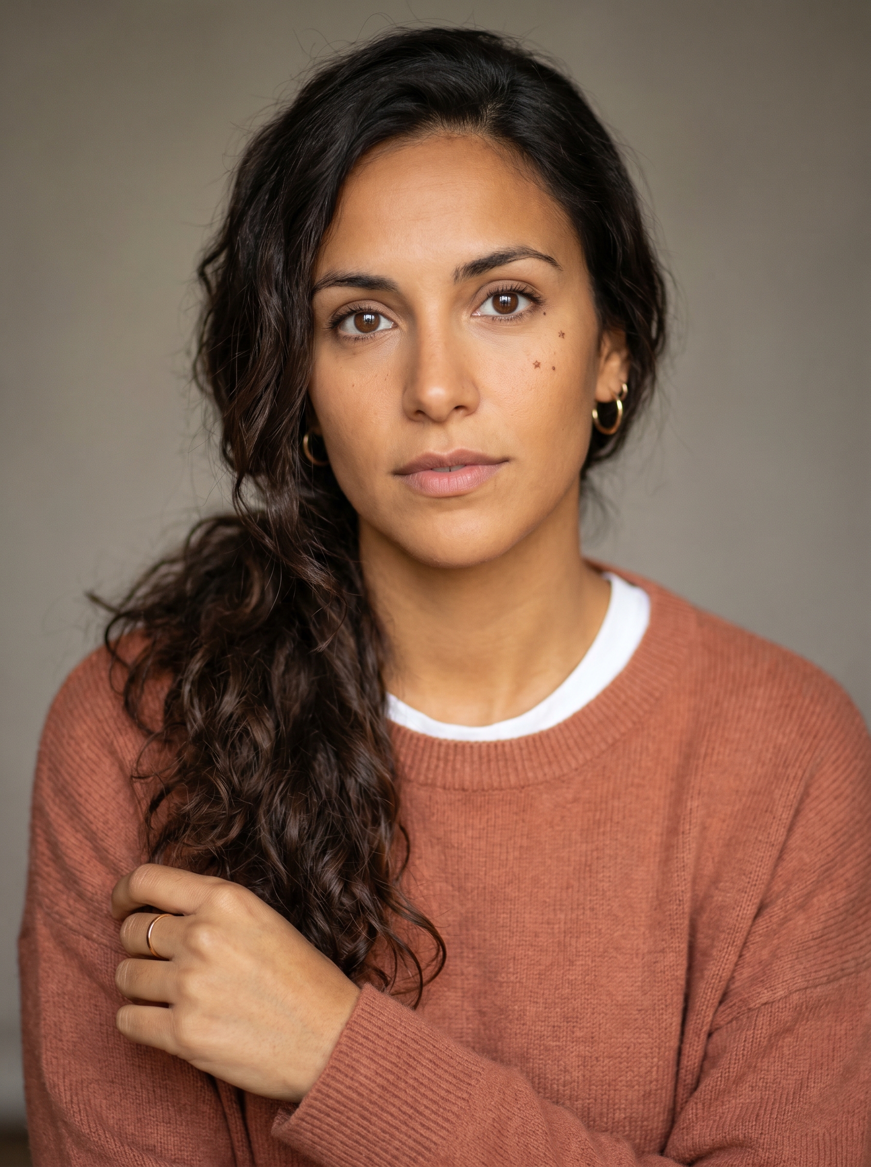 A Spanish woman in her late 20s, Seville-born, with the warm, kinetic attentiveness of someone who sees movement and flow where others see steps. Medium height, poised, with an easy, expressive physicality. Warm golden-brown skin with a healthy glow. Dark, near-black hair, thick and wavy, worn long and loosely gathered over one shoulder. Large dark-brown eyes, bright and quick, under naturally strong brows. An oval face with full cheeks and a ready, generous smile she does not suppress. A faint scattering of freckles high on the cheeks. She wears a soft terracotta knit with the sleeves pushed up over a fine white tee, small gold hoop earrings, the wardrobe of someone warm and current without trying. Distinguishing feature: a small star-shaped freckle beneath the right eye, and a thin gold ring worn on the index finger.