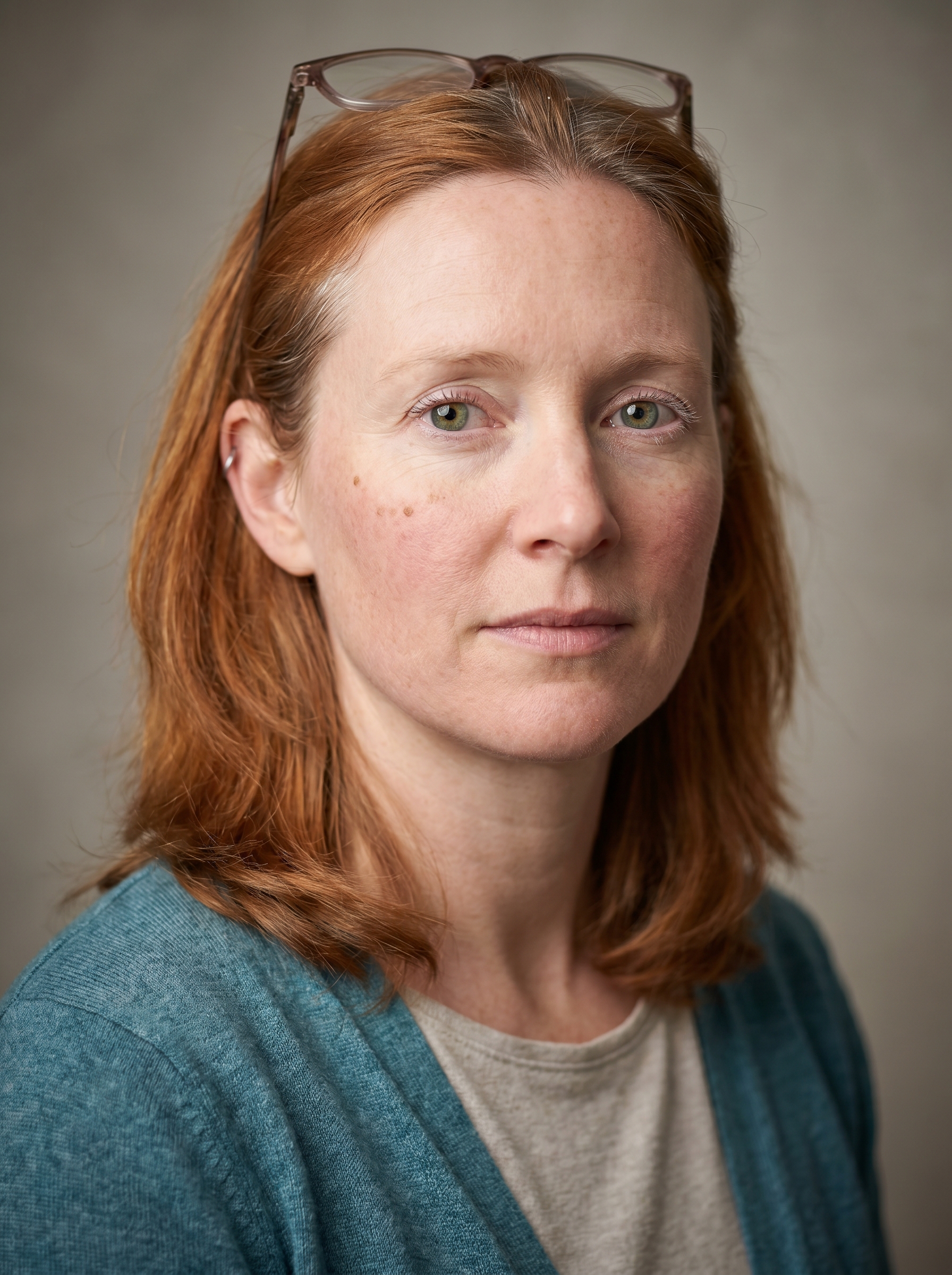An Irish woman in her early 40s, Cork-born, with the grounded, observant practicality of someone who has surveyed every corner of how the work actually gets done. Medium build, sturdy and relaxed in her stance. Fair, warm-toned skin with light freckling across the nose and a faint windburn at the cheeks. Coppery-auburn hair, thick and shoulder-length, worn half-pinned back, a few greys catching the light. Clear green eyes, frank and friendly, with pale lashes and soft laugh lines. A round, open face with a slightly upturned nose. She wears a heathered teal cardigan over a simple oatmeal top, reading glasses pushed up into her hair, a canvas field-notebook in hand, the wardrobe of someone who works in the field, not the boardroom. Distinguishing feature: a small constellation of three freckles on the left cheekbone, and a thin silver hoop in the upper left ear.